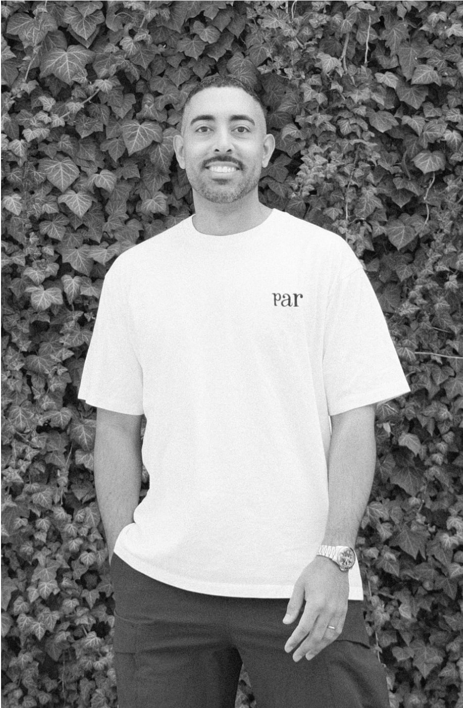 Black and white headshot. White shirt, in front of a green vine wall.